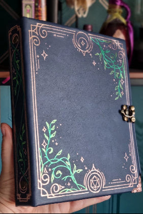 a dark navy blue notebook with an ornate gold border and green botanical illustrations, featuring a small metal clasp on the side.